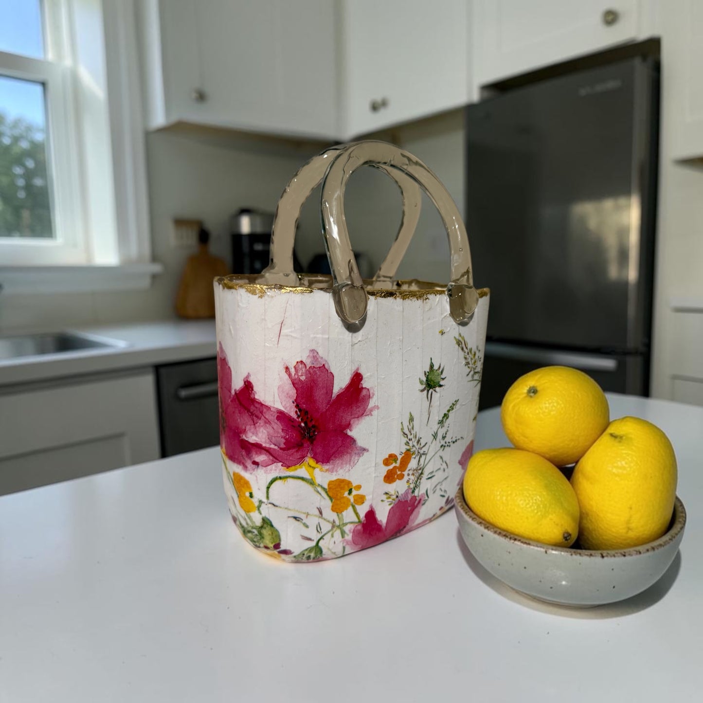 A glass purse vase with a floral design sitting on a kitchen counter next to a bowl of lemons.