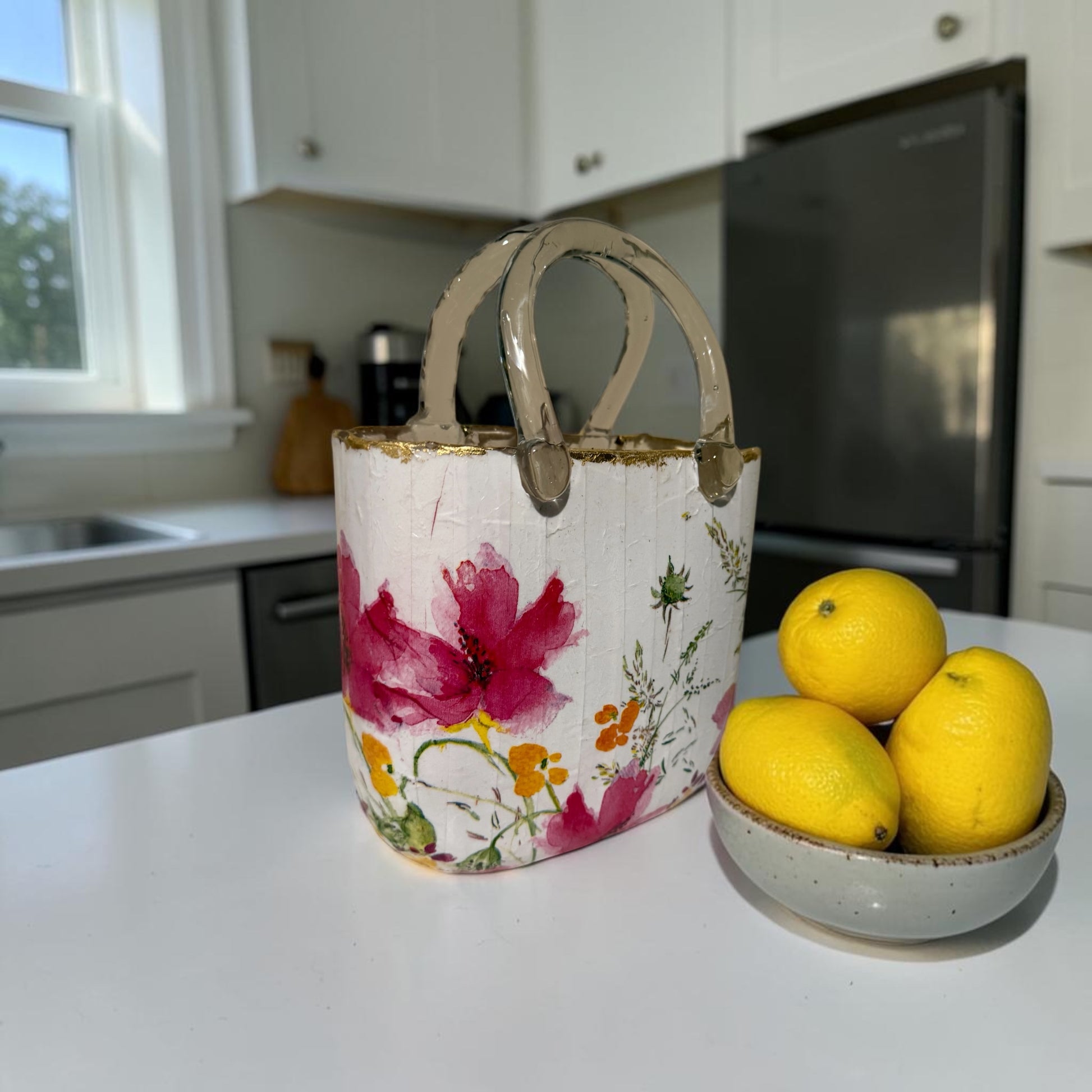 A glass purse vase with a floral design sitting on a kitchen counter next to a bowl of lemons.