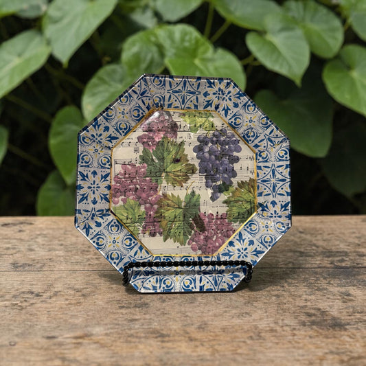 Decorative plate with blue pattern on the edge and a grape design  over sheet music on a concrete surface with green leaves in the background