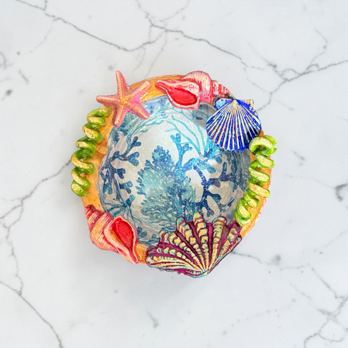 A handmade seashell and coral-themed decorative bowl with a blue and white coral print center, surrounded by colorful clay seashells, a starfish, and green coiled ribbon.